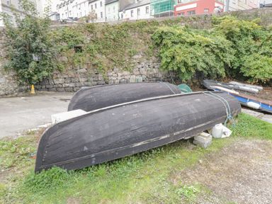 Two black upturned boats on grass near a stone wall with greenery and houses in the background at Sea Watch House in Roundstone County Galway