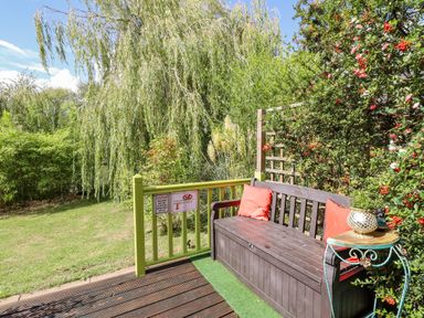 An outdoor wooden bench with orange cushions next to a small table on a deck overlooking a garden with trees and plants at Lakeside Lodge Retreat in Tattershall