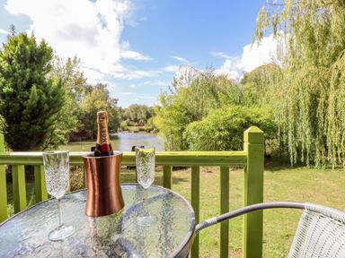 A glass table with a champagne bottle in an ice bucket and two glasses on a balcony overlooking a river and green trees at Lakeside Lodge Retreat in Tattershall