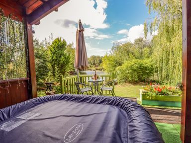 A covered patio with a covered hot tub and a small table with two chairs and an umbrella overlooking a garden and lake at Lakeside Lodge Retreat in Tattershall