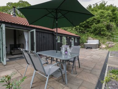 A patio area with a table and chairs under a large umbrella and a hot tub near a building with an open door at Bottom Lodge in Penhow
