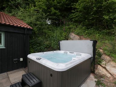 An outdoor hot tub with water and steps next to a black wooden building with a red roof at Bottom Lodge in Penhow