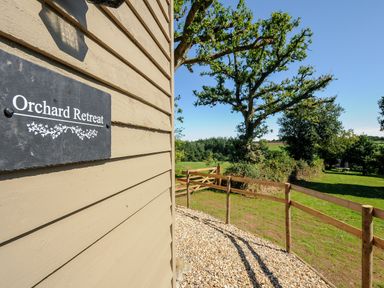 The side of a building with a sign reading Orchard Retreat and a fenced grassy area with trees at Orchard Retreat East Worlington near Witheridge