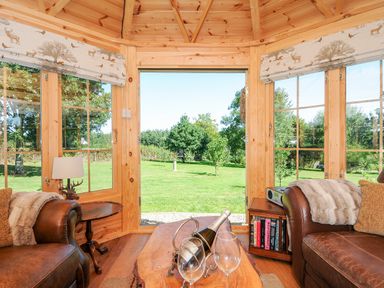 A wooden room with two leather chairs a wooden table with wine and glasses and large windows overlooking a green garden at Orchard Retreat in East Worlington near Witheridge
