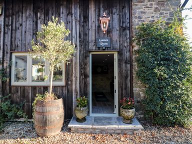 An entrance to a games room with a wooden exterior wall a wooden barrel planter with a tree and two stone planters with flowers at Orchard Retreat in East Worlington near Witheridge