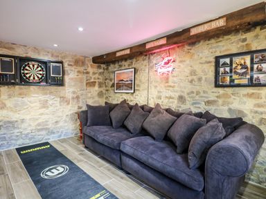 A room with a dark grey sofa darts board and framed pictures on stone walls at Orchard Retreat in East Worlington near Witheridge