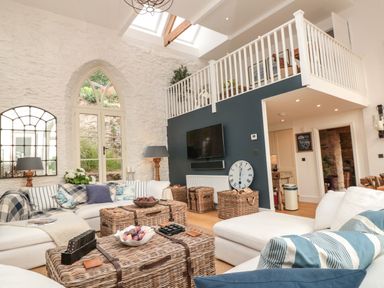 A living room with white sofas, wicker tables, large arched window, and a loft area at The Old Chapel in Dittisham