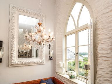 A dining nook with a chandelier a large ornate mirror an arched window with shutters and candles on the sill at The Old Chapel in Dittisham