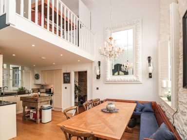 A kitchen and dining area with wooden table and corner bench seating under a chandelier and balcony at The Old Chapel in Dittisham
