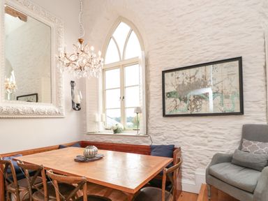 A dining area with a wooden table and chairs under a chandelier next to an arched window with pillows and a framed map on the wall at The Old Chapel in Dittisham