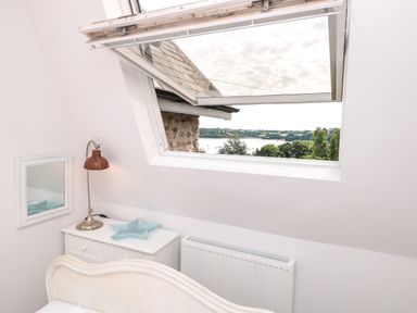 A bedroom corner with a bedside table lamp and open roof window overlooking trees and water at The Old Chapel in Dittisham