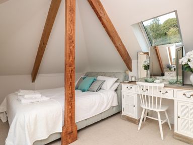 A bedroom with a bed and white bedding and a vanity table with a chair under a slanted ceiling with wooden beams and a window at The Old Chapel in Dittisham