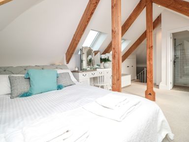 A bedroom with a bed pillows a dressing table with mirror and flowers exposed wooden beams and an en suite bathroom at The Old Chapel in Dittisham
