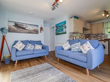 A living room with two blue sofas and cushions with a kitchen area in the background at Woodside in Rosecraddoc near St Cleer