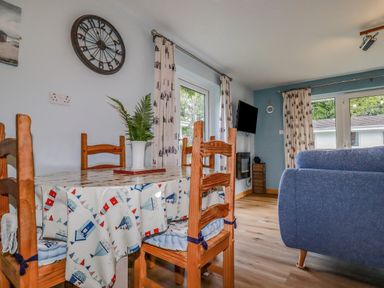 A dining area with wooden chairs and table with a patterned tablecloth and a living room with blue sofas and a wall-mounted TV at Woodside in Rosecraddoc near St Cleer