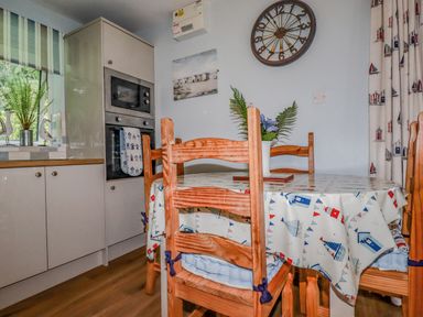 A kitchen with wooden chairs around a table covered with a patterned tablecloth open shelving and a wall clock at Woodside Rosecraddoc near St Cleer