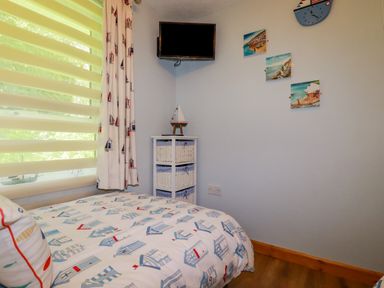 A bedroom with a bed near a window with blinds and curtains a small white storage unit a wall mounted television and three pictures on the wall at Woodside Rosecraddoc near St Cleer