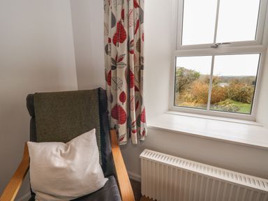A corner with a chair with cushions next to a window with patterned curtains and a radiator below at Ceunant near Newport