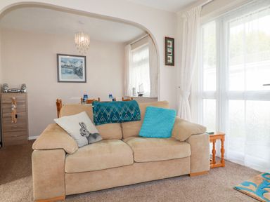 A living room with a beige sofa and cushions next to a wooden side table and large window at Smugglers in Liskeard near St Cleer