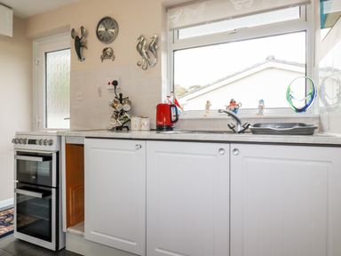 A kitchen with a stove white cabinets a sink and window with decorative items on the sill at Smugglers in Liskeard near St Cleer