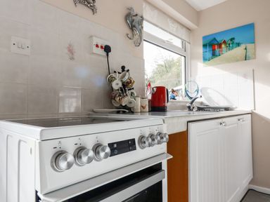 A kitchen countertop with a stove a red kettle mugs on a rack and a window with decorations at Smugglers in Liskeard near St Cleer