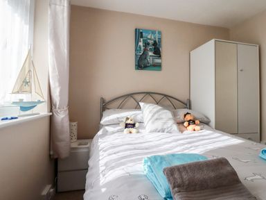 A bedroom with a bed has two stuffed toys and folded towels with a wardrobe and a bedside table near window with curtains at Smugglers in Liskeard near St Cleer