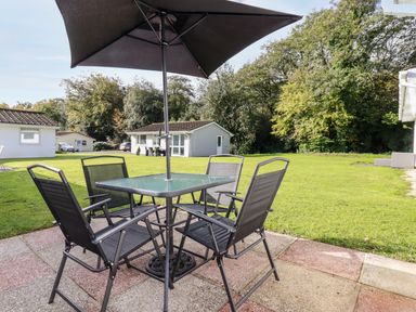 An outdoor patio area with a glass table four black chairs and a large umbrella on a lawn with small houses and trees at Smugglers in Liskeard near St Cleer