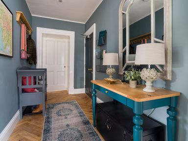 An entryway with a blue bench, a wooden table with lamps and plants, a large mirror, a coat rack, and a rug on a wooden floor at Wansfell at Lipwood House in Windermere
