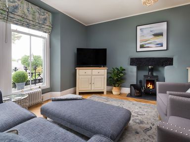 A living room with a blue sofa and ottoman a television on a cabinet a fireplace and a window with a potted plant at Wansfell at Lipwood House Windermere