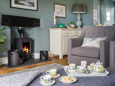 A living room with a burning stove a gray armchair and a tea set on a table at Wansfell at Lipwood House in Windermere