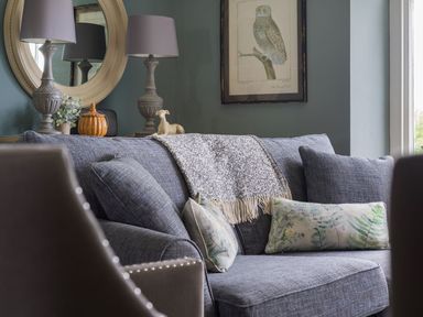 A living room with a blue sofa pillows a blanket two lamps on a table a round mirror and a framed owl picture on the wall at Wansfell at Lipwood House in Windermere