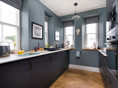 A kitchen with blue walls dark cabinets wooden floor and three windows with gray blinds at Wansfell at Lipwood House in Windermere