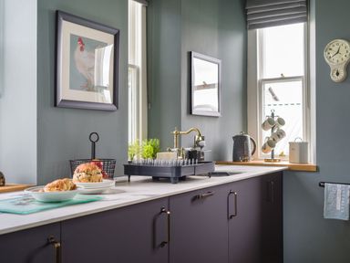 A kitchen countertop with baked goods on plates a wire basket with apples plants a gold faucet and a kettle near a window Wansfell at Lipwood House Windermere