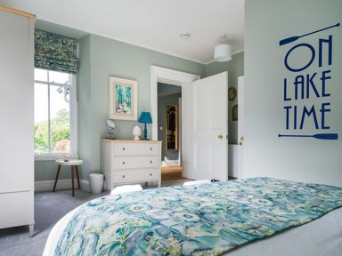 A bedroom with a bed covered in blue and green bedding a dresser a small side table and a window with patterned curtains at Wansfell at Lipwood House in Windermere