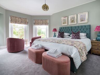 A bedroom with a teal upholstered bed with pillows and a floral bed runner two pink ottomans with towels a pair of pink chairs near windows with floral blinds and wooden bedside tables with lamps Wansfell at Lipwood House in Windermere