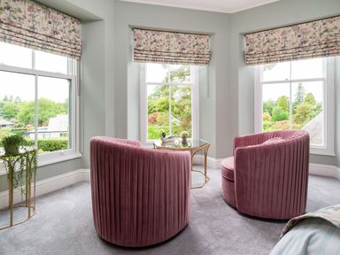 A sitting area with two pink chairs and a glass table by three windows with floral blinds at Wansfell at Lipwood House in Windermere