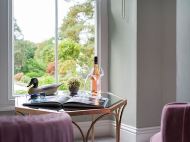 A corner table with a book sunglasses a duck figurine a small plant a wine bottle and two glasses by a window at Wansfell at Lipwood House in Windermere