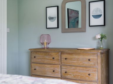 A wooden chest of drawers with a glass lamp and a vase with flowers below two framed pictures and a mirror at Wansfell at Lipwood House in Windermere