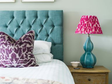 A bedroom with a blue tufted headboard purple patterned pillows a blue lamp with a pink shade on a wooden nightstand at Wansfell at Lipwood House in Windermere