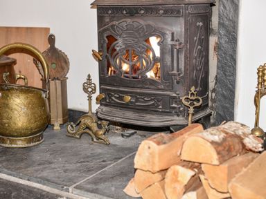 A close-up of a wood-burning stove with fire inside with firewood stacked nearby and brass fireplace tools at Naunton Cottage in Lyme Regis