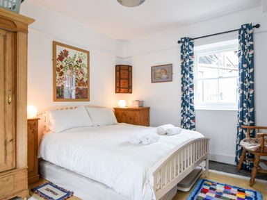 A bedroom with a white bed and rolled towels next to wooden furniture and a window with blue patterned curtains at Naunton Cottage in Lyme Regis