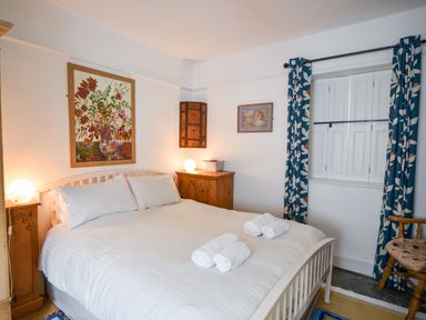 A bedroom with a white bed and towels on it bedside tables with lamps paintings on the walls and blue curtains at Naunton Cottage in Lyme Regis