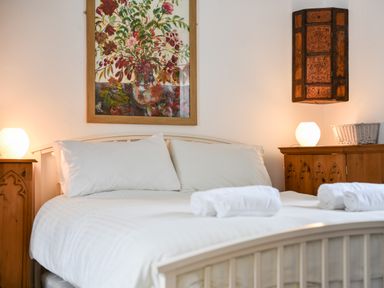 A bedroom with a bed with white linens and pillows two wooden bedside cabinets with lamps and a wall painting at Naunton Cottage in Lyme Regis