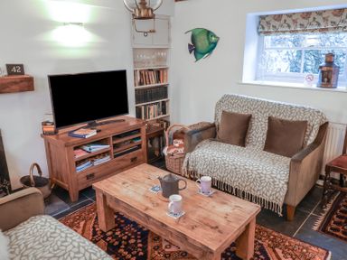 A living room with a wooden coffee table mugs teapot sofas a television on a wooden stand and a window at Naunton Cottage in Lyme Regis