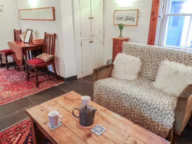 A living room with a wooden coffee table with a teapot and cups a patterned sofa with cushions wooden chairs and a table against the wall at Naunton Cottage in Lyme Regis
