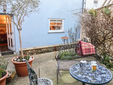 A small courtyard with a metal table and chairs a bench with a blanket potted plants and a blue wall with a window at Naunton Cottage in Lyme Regis