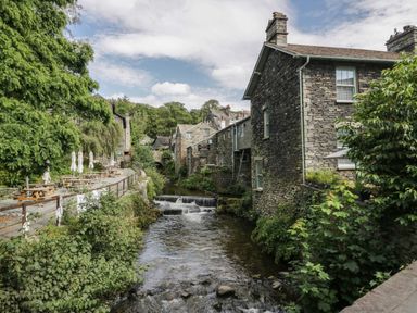 A river flowing between stone buildings and trees with picnic tables along the river at Rose Cottage Ambleside