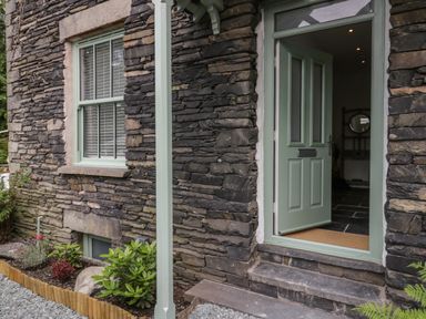 A stone exterior wall with a green framed window and an open green door showing a glimpse of an interior with tiled floor at Rose Cottage in Ambleside