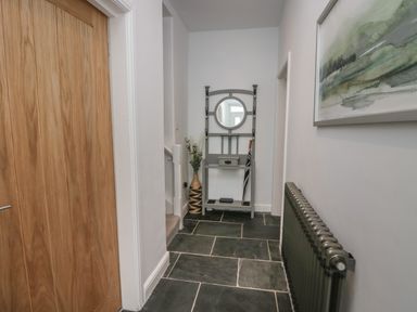 A hallway with a wooden door to the left a radiator on the right and a small grey hall stand with a round mirror and umbrella at Rose Cottage Ambleside