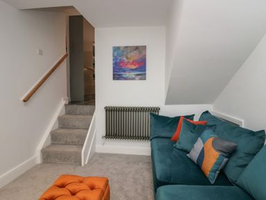 A small living room with a teal sofa and colorful pillows near carpeted stairs and a wall radiator with a painting above at Rose Cottage in Ambleside